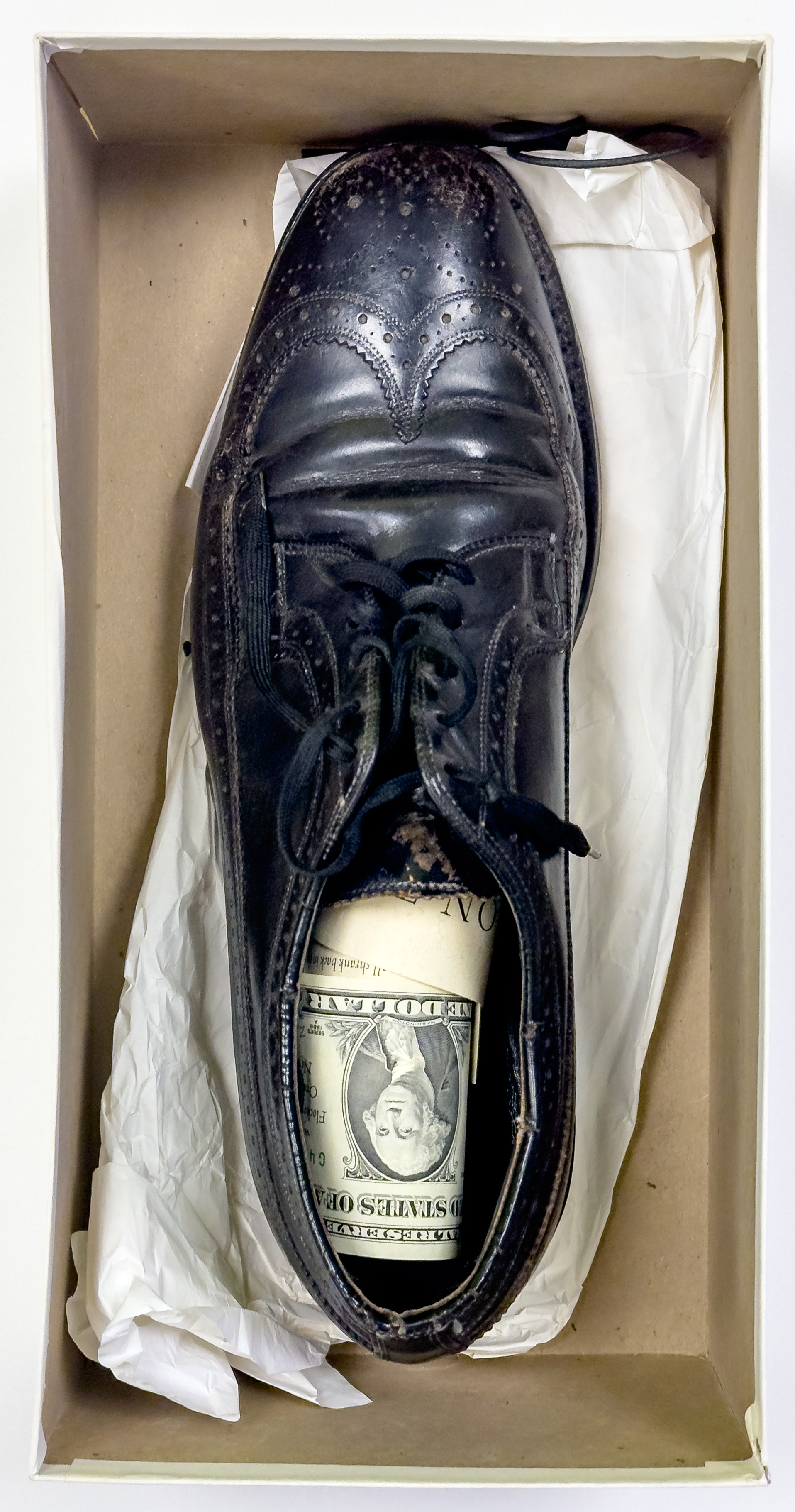 An image of a shoe box with a black dress shoe with a dollar bill inside of it