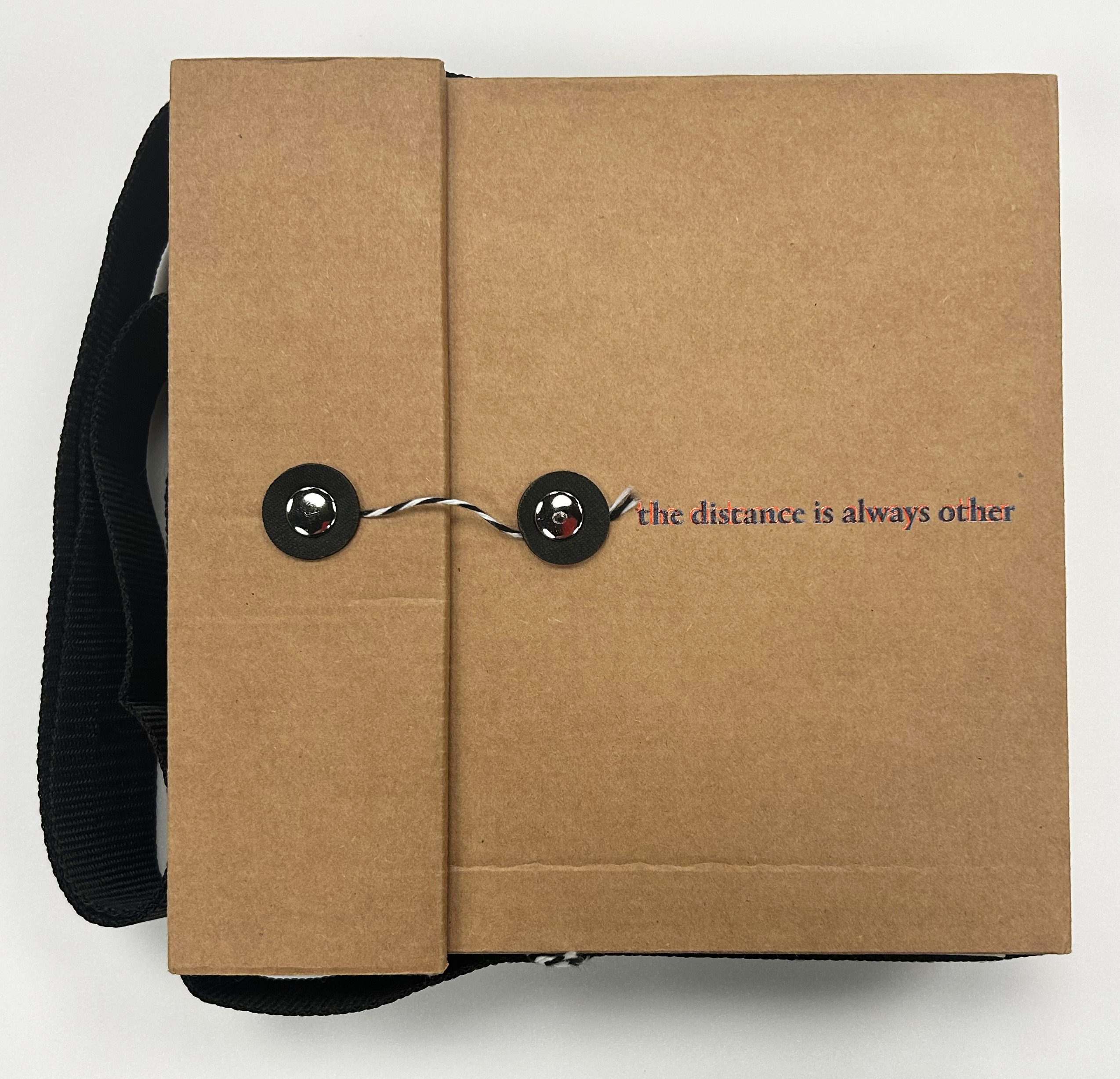 A brown cardboard button string closure book with the words, "The Distance Is Always the Other," in black with red behind it. 